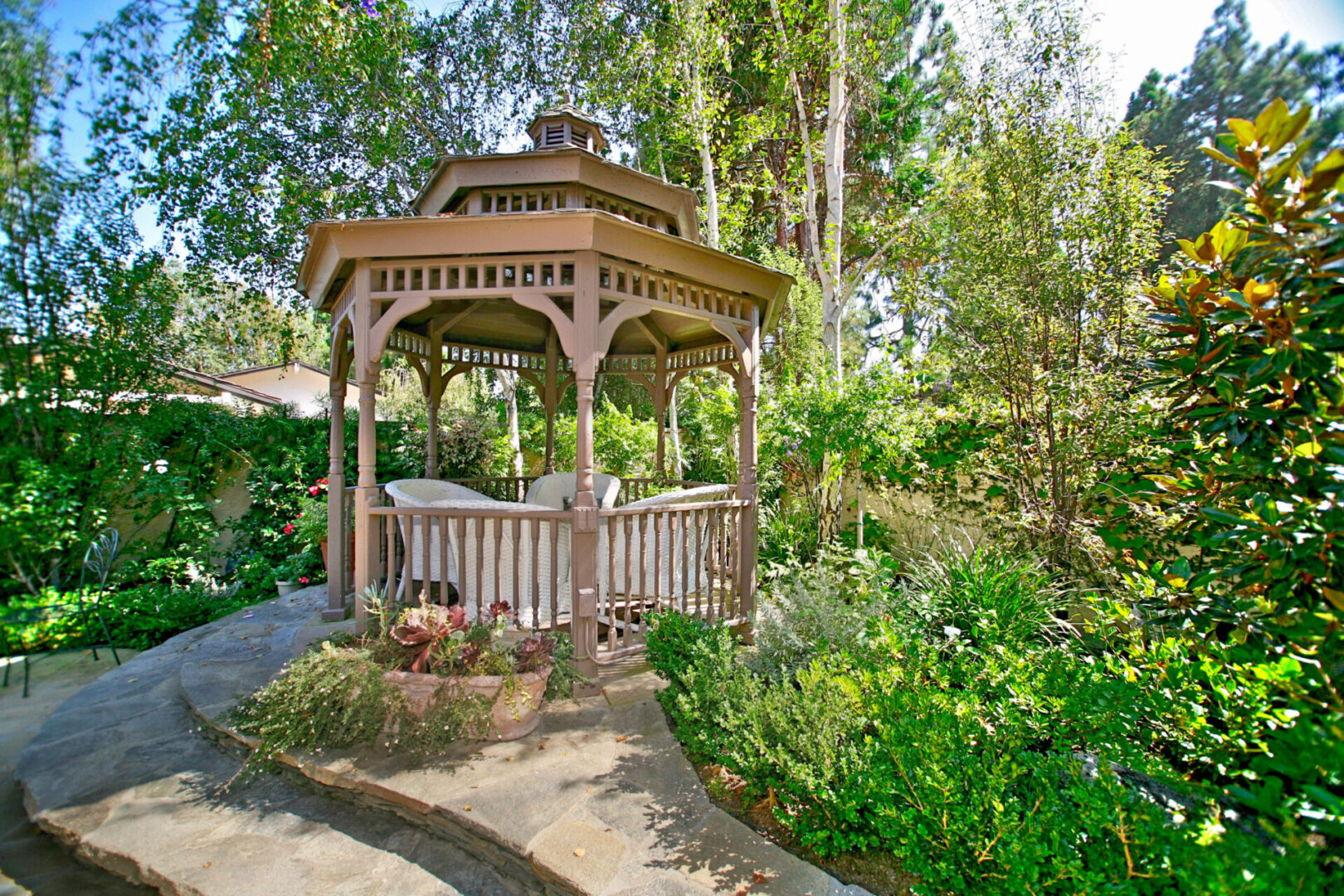 A wooden gazebo surrounded by trees and flowers.